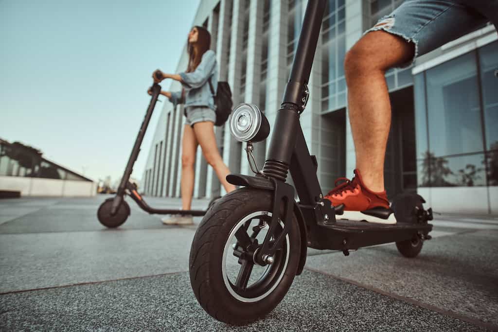 Ein Junges Paar fährt mit E-Scooter unter einem sonnigen Himmel. Im Hintergrund ist ein Gebäude mit Architektonischen modernen Stil und großen Glasfenster zu sehen.