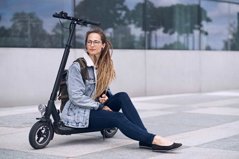 Eine Junge Frau mit Brille und langen aschblonden Rasta Haaren und einer Jeans Jacke in hellem Blau mit dunklblauer Enganliegender Jeans und schwarzen Ballarinas sitz gemütlich auf einen E-Scooter in Schwarz.